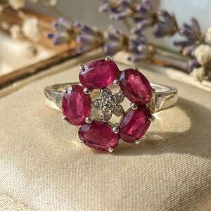 May include: A silver ring with a flower-shaped design, showcasing six oval, deep red gemstones encircling a central cluster of smaller, clear stones. The ring is set on cream fabric, with a wooden box and floral elements in the background.