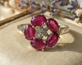 May include: A silver ring featuring a floral design with six oval-cut, deep red gemstones surrounding a cluster of smaller, clear stones. The ring is set against a cream-colored fabric, with a wooden box and floral accents in the background.