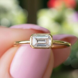 May include: A gold ring featuring a rectangular emerald-cut diamond, set within a bezel. The ring is held up against a blurred green backdrop, with a focus on the diamond's clarity.