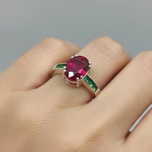 Peut inclure: Une bague en argent ornée d'un grand rubis ovale en pierre centrale. La bague est flanquée de deux pierres vertes taille émeraude de chaque côté. La bague est portée sur une main.