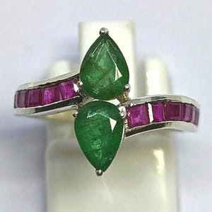 May include: A silver ring featuring two pear-shaped green emeralds and a band of small, rectangular, red rubies. The ring is displayed on a white stand against a white background. The gemstones are the focal point of the ring.