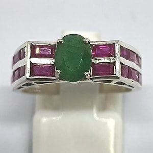 Peut inclure: Bague en argent ornée d'une émeraude verte ovale au centre, encadrée de pierres rouges rectangulaires. La bague est présentée sur un présentoir blanc, mettant en valeur les couleurs vives et le design détaillé du bijou.