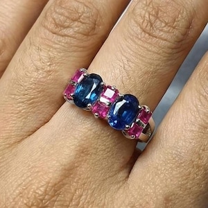 May include: A silver ring with three oval blue gemstones and two rows of rectangular pink gemstones. The ring is displayed on a hand, highlighting the contrast between the blue and pink gemstones.