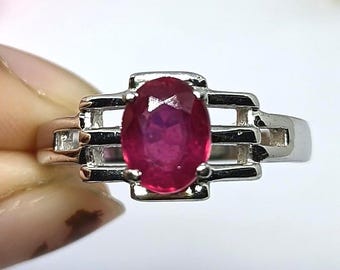 May include: A silver ring featuring a large, oval-cut ruby gemstone. The ruby is a deep, vibrant red color. The ring has a geometric design with rectangular accents on either side of the ruby. The ring is set against a blurred background of flowers.