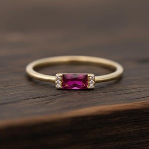 Puede incluir: Un anillo de oro con una piedra preciosa rectangular de color magenta en el centro. Pequeñas piedras claras flanquean la piedra principal. El anillo se encuentra sobre una superficie de madera oscura, resaltando el diseño de la joya.