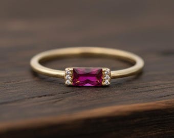 Pode incluir: Um anel de ouro com uma pedra preciosa retangular magenta no centro. Pequenas pedras transparentes ladeiam a pedra principal. O anel está sobre uma superfície de madeira escura, realçando o design da joia.