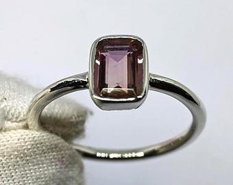 May include: A silver ring featuring a rectangular, light purple gemstone. The gemstone is set in a bezel setting, and the band is thin and polished. The ring is held by a gloved hand against a soft, blurred background.