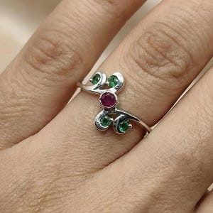 Puede incluir: Un anillo de plata con un diseño en espiral, con una gema roja central y varias gemas verdes. El anillo está colocado en un dedo, mostrando sus delicados detalles y colores vibrantes. El diseño del anillo se asemeja a hojas.
