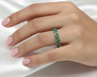 Könnte beinhalten: Ein silberner Ring mit zwei Reihen kleiner, runder, smaragdgrüner Edelsteine. Der Ring wird an einem Finger getragen, der auf einer weißen, seidigen Oberfläche ruht. Die eng aneinander liegenden Edelsteine bilden ein durchgehendes Farbband.