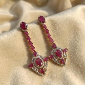 Könnte beinhalten: Ein Paar goldfarbene Ohrhänger mit ovalen und runden rubinroten Edelsteinen. Die Ohrringe haben ein detailliertes Design mit kleineren Steinen, die die größeren rubinroten Edelsteine umgeben. Die Ohrringe liegen auf einer hellbeige Oberfläche.