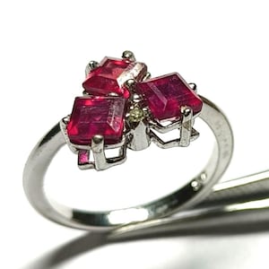 Peut inclure: Une bague en argent ornée de trois pierres précieuses rouges rectangulaires vibrantes, disposées en motif floral. La bague a un anneau fin, et les pierres sont serties dans un sertissage détaillé et orné. La bague est présentée sur fond blanc.