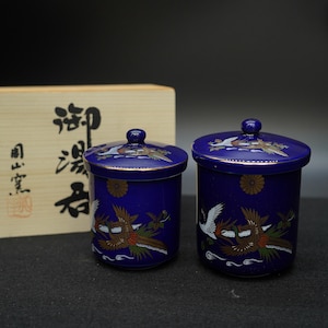 以下が含まれることがあります： コバルトブルーの蓋付きセラミックジャー2個。各ジャーは金色の縁取りが施され、鶴と花の模様で装飾されています。ジャーは円筒形で、蓋の上部に小さなノブが付いています。