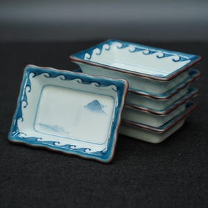以下が含まれることがあります： 小さくて長方形の陶器の小皿が6枚重ねられています。各皿は白い内側で、青い山と波のデザインが施されています。縁は青色で波模様があり、茶色の縁取りがあります。皿は暗い表面に積み重ねられています。