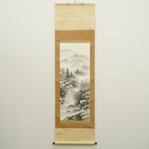 Peut inclure: Peinture japonaise sur rouleau représentant un paysage de montagne brumeux avec des arbres et une rivière. L'œuvre est en nuances de gris et de blanc, encadrée par une bordure marron et du papier beige. Le rouleau est suspendu à une baguette en bois.