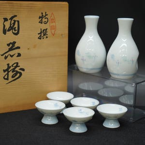以下が含まれることがあります： 白磁の酒器セット。徳利2本と盃4個。ボトルとカップには青い花柄と底に青い帯があります。背景には漢字の書かれた木箱があります。