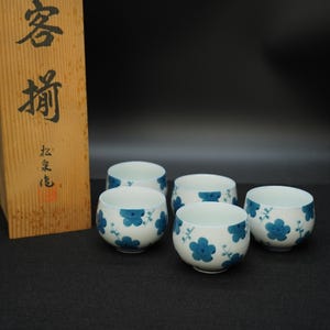 以下が含まれることがあります： 青い花の模様が施された、白い陶器の湯呑み5個セット。湯呑みはグループで配置され、左側には木箱があります。木箱には日本語の文字が書かれています。