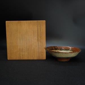 May include: A handmade ceramic bowl with a wide rim and a footed base. The bowl features a mix of colors, including brown, green, and beige. A wooden box is next to the bowl.