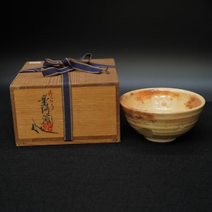 May include: A ceramic bowl with a light brown and orange glaze sits next to a wooden box tied with a blue ribbon. The bowl has a rounded shape with a textured surface. The box has Japanese characters and artwork.