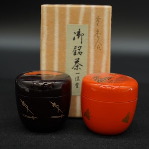 以下が含まれることがあります： 蓋付きの小さな丸い漆器容器が2つ。1つは金色の線が入ったダークブラウン、もう1つは黒と金の装飾が施された鮮やかなオレンジ色で、縦縞模様と日本語の書が書かれた長方形のベージュ色の箱の前に置かれています。
