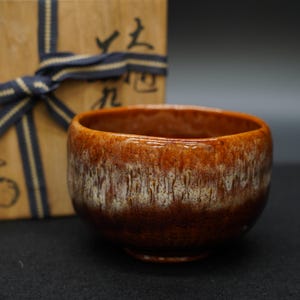 Peut inclure: Un bol à thé en céramique avec une finition brillante et texturée. Le bol présente un dégradé de teintes marron et blanches. Une boîte en bois avec des caractères japonais et un ruban bleu est en arrière-plan.