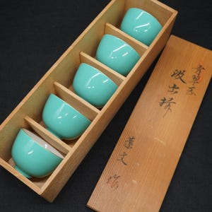 以下が含まれることがあります： 木製の箱に収められた、金色の縁取りが施されたターコイズ色の陶器のティーカップ5客セット。箱には、日本の書道が施されたスライド式の蓋が付いています。