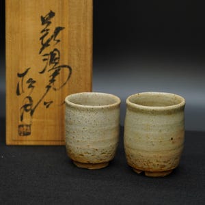 以下が含まれることがあります： 薄いベージュ色の、斑点のある小さな陶器のカップが2つ。カップは円筒形で、縁は丸く、小さな台座の上にあります。背景には、日本の書道が書かれた木箱があります。