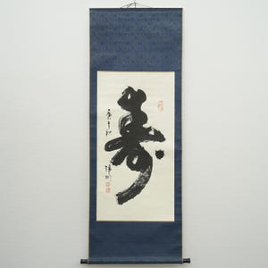 May include: A traditional Japanese scroll painting with black calligraphy on a white background. The scroll features a large, stylized character and smaller vertical text. The top and bottom are dark blue with a decorative pattern.