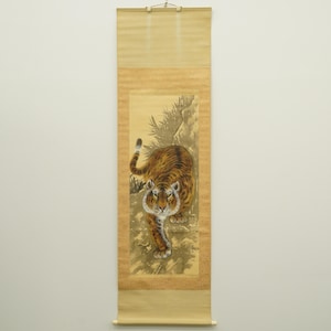 May include: A vintage Asian scroll painting featuring a detailed depiction of a tiger. The tiger, rendered in shades of brown and orange with black stripes, is set against a backdrop of rocks and foliage. The scroll is framed in a light tan color.