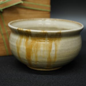 以下が含まれることがあります： 丸みを帯びた形の、手作りの陶器のボウル。オフホワイトで、茶色と黄色の縞模様があります。釉薬は光沢のある仕上がりです。デザインは陶器作品を示唆しています。