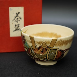 Puede incluir: Cuenco de té de cerámica color crema con un diseño detallado de casco de guerrero en tonos marrones, verdes y rojos. El cuenco se presenta con una caja roja, y el texto de la caja dice "Cuenco de té".