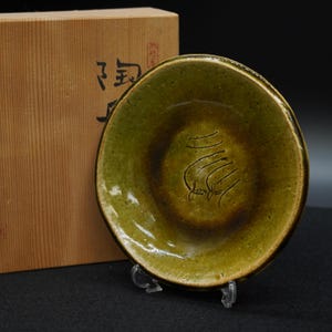 以下が含まれることがあります： 手作りのオリーブグリーンのセラミックプレート。中央は濃い茶色で、花の模様が彫られています。透明なスタンドに飾られ、横には日本語の文字が書かれた木箱があります。