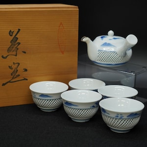 Puede incluir: Un juego de té de cerámica blanca con diseños de montañas azules, que incluye una tetera y cinco tazas. Las tazas y la tetera tienen un patrón tejido alrededor de la base. El juego se exhibe con una caja de madera.