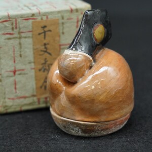 May include: A small, glazed ceramic figurine featuring a rounded, light brown body with a fine crackle finish. It has a dark, glossy black head section with a circular yellow accent. The figurine sits in front of a light-colored box with red lines.