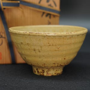 May include: A beige ceramic tea bowl with a textured, ribbed exterior and speckled brown accents. The bowl features a wide, open rim and a sturdy footed base. It is presented in front of a wooden storage box tied with a dark blue ribbon.
