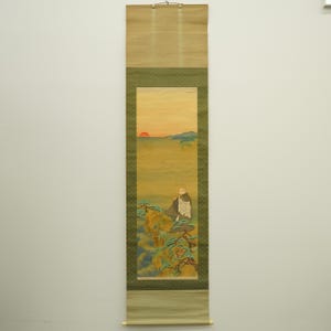 May include: A traditional Japanese scroll painting depicting a figure seated on a hillside, gazing at a sunset. The artwork features a warm color palette with shades of yellow, orange, and green. The scroll is framed with a green border and has a wooden hanging rod.