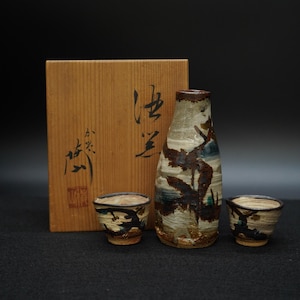 May include: A ceramic sake set featuring a bottle and two cups, with a wooden box in the background. The bottle and cups have a textured, earthy design with brown, blue, and beige tones. The wooden box has Japanese calligraphy.