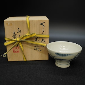 May include: A white ceramic bowl with blue painted designs, next to a wooden box tied with a yellow ribbon. The bowl has a pedestal base and a rounded rim. The box has Japanese text and a red stamp.