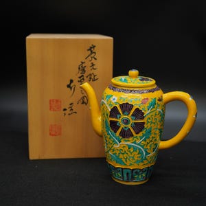 以下が含まれることがあります： 紫とティール色の花柄が施された黄色のセラミックティーポット。 ティーポットにはお揃いの蓋とハンドルが付いています。 ティーポットの後ろには、日本語の文字が入った木箱があります。