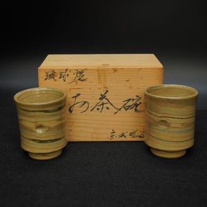 Könnte beinhalten: Zwei beigefarbene Keramik-Teetassen mit horizontalen Streifen und eine Holzkiste mit japanischen Schriftzeichen. Die Teetassen haben eine zylindrische Form mit leicht ausgestelltem Boden. Die Kiste ist hellbraun mit schwarzer Kalligraphie.