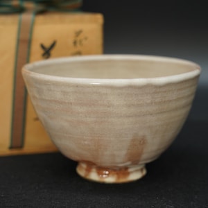 May include: A ceramic tea bowl with a light beige, textured glaze and subtle brown drip patterns near the base. The bowl features a slightly uneven rim and a raised foot. Behind the bowl sits a tan wooden storage box with black Japanese calligraphy.