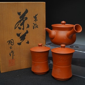 Puede incluir: Un juego de té de terracota con una tetera y dos tazas con tapa, junto a una caja de madera con caligrafía japonesa. La tetera tiene un cuerpo redondeado y un pico curvo. Las tazas son cilíndricas con diseños florales en relieve.