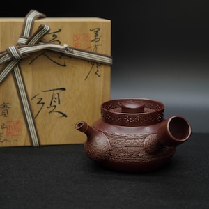 May include: A reddish-brown ceramic teapot with a textured surface and a decorative lid. The teapot has a spout and a handle. A wooden box with calligraphy and a ribbon is in the background.