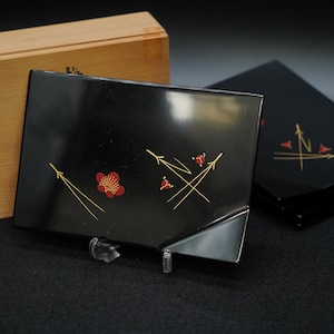 以下が含まれることがあります： 赤と金のフローラルデザインが施された黒漆塗りのサービングトレイ。トレイは光沢のある仕上がりで、透明なスタンドに展示されています。背景には木箱があります。トレイはセットの一部です。