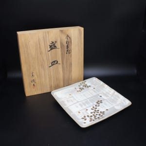 Könnte beinhalten: Ein quadratischer, cremefarbener Keramikteller mit floralem Design und eine Holzkiste. Der Teller hat braune Blumenmuster und eine strukturierte Oberfläche. Die Holzkiste ist mit japanischen Schriftzeichen bedruckt.