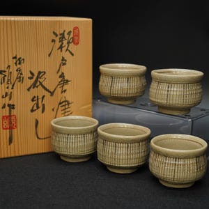 May include: A set of five beige ceramic teacups with a textured, striped design. The cups are arranged in front of a wooden box with Japanese calligraphy. The cups are approximately 3 inches in diameter and 2 inches tall.