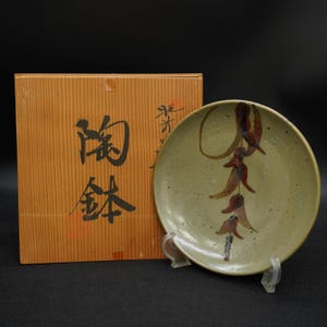 Puede incluir: Un plato de cerámica con una superficie moteada verde claro, con un diseño de chiles rojos colgantes. El plato se exhibe sobre un soporte transparente, junto a una caja de madera con caracteres japoneses.