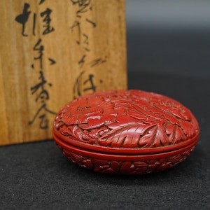 May include: A round, cinnabar-red lacquer box featuring intricate, deeply carved floral and leaf motifs across its lid and sides. The container is presented alongside a light-colored wooden storage box marked with black Japanese calligraphy.