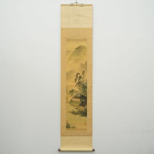 May include: A vertical scroll painting depicting a landscape with mountains, trees, and a small building. The artwork features a muted color palette with shades of yellow, green, and brown. The scroll is framed with a patterned border and has wooden dowels at the top and bottom.