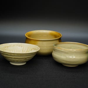 以下が含まれることがあります： 暗い表面に、さまざまなサイズとベージュと茶色の色合いの3つのセラミックボウルが配置されています。 ボウルは素朴で手作りの外観で、目に見える質感と微妙な色のバリエーションがあります。 ボウルはおそらく、サービングまたはディスプレイ用です。