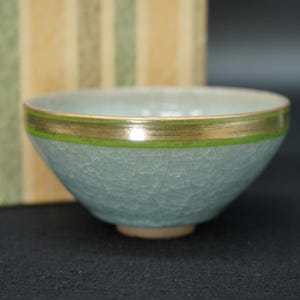 以下が含まれることがあります： 淡い青緑色の貫入釉仕上げが施された陶器製の茶碗。縁には鮮やかな緑色の帯とメタリックゴールドの縁取りが施されています。丸みを帯びた形状で、小さな円形の高台の上に置かれており、繊細な質感が特徴です。